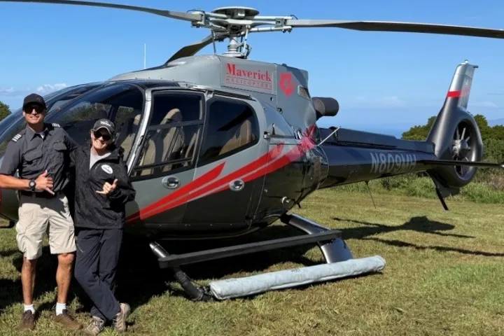 Two people posing in front of a parked helicopter on grass.